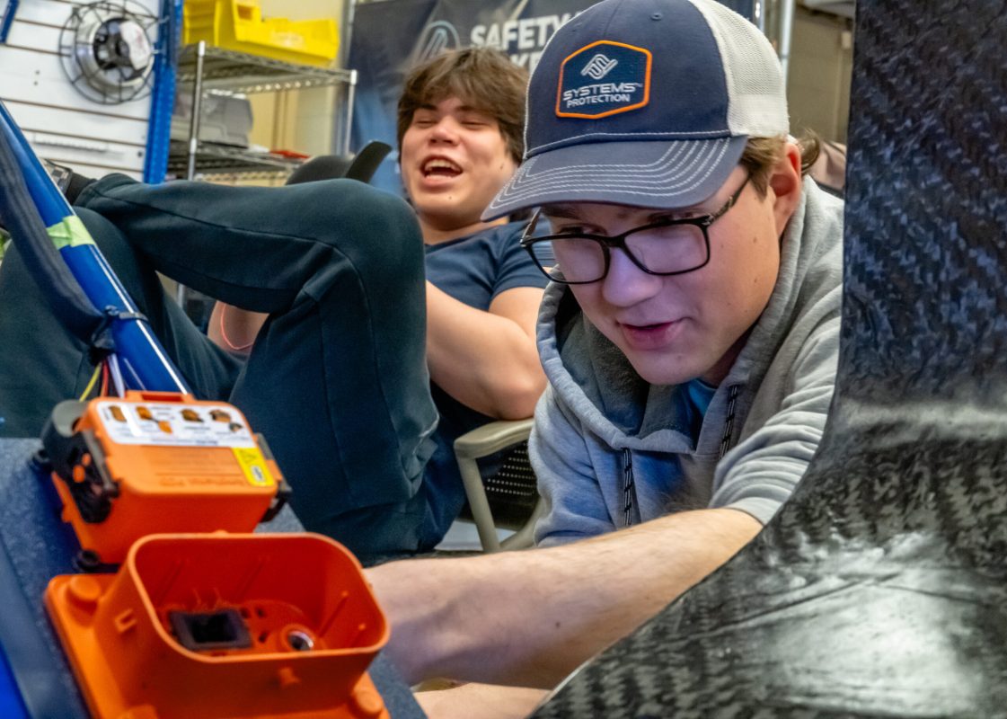 Nittany Motorsports team member works on the team's car while another sits close joking with others in the shop.